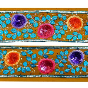 Op de afbeelding: Een gouden stoffen bies met een herhalend patroon van kleurrijke geborduurde bloemen en bladeren. De bloemen zijn gemaakt van pailletten en hebben een verscheidenheid aan kleuren, waaronder roze, oranje, paars en blauw.