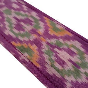 May include: Purple fabric with a green and orange ikat pattern. The fabric has a stitched edge.