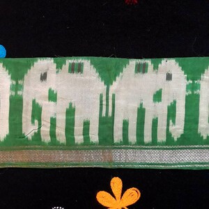 Può includere: Primo piano di un tessuto verde e bianco con un motivo di elefanti. Il tessuto è bordato con un bordo argentato. Il tessuto è su uno sfondo nero con un piccolo disegno di fiore arancione.