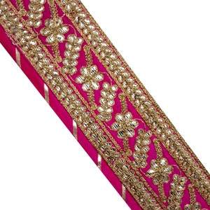 Può includere: Primo piano di una vibrante striscia di tessuto fucsia con intricati ricami dorati. Il design presenta motivi floreali e a foglie con abbellimenti scintillanti. Sottili strisce argentate corrono lungo il bordo del tessuto.