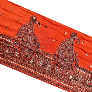 May include: Orange fabric with intricate embroidery featuring paisley designs and sparkling embellishments. The fabric has a textured, slightly wrinkled appearance.