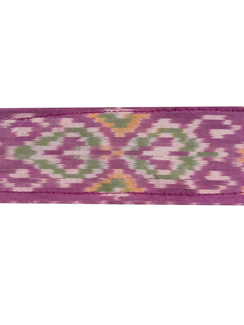 May include: A close-up of a purple fabric with a green and white ikat pattern. The fabric has a pink stitched edge.