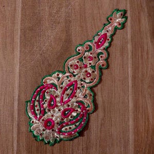 Puede incluir: Una aplicación bordada de color verde, rosa y dorado con un diseño floral. La aplicación está decorada con lentejuelas y cuentas.