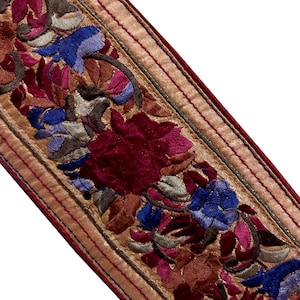May include: Close-up of a decorative trim with an embroidered floral design. The trim has a gold satin base with a burgundy border and is adorned with flowers in shades of burgundy, blue, pink, and brown.