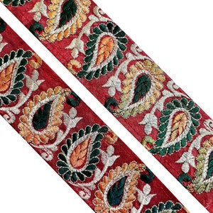 May include: Two strips of red fabric with embroidered paisley designs. The paisley elements feature green, orange, and yellow details, outlined with white thread. The fabric has a textured appearance, and the design is symmetrical.