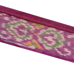 May include: A close-up of a purple fabric with a green and white ikat pattern. The fabric has a pink border.
