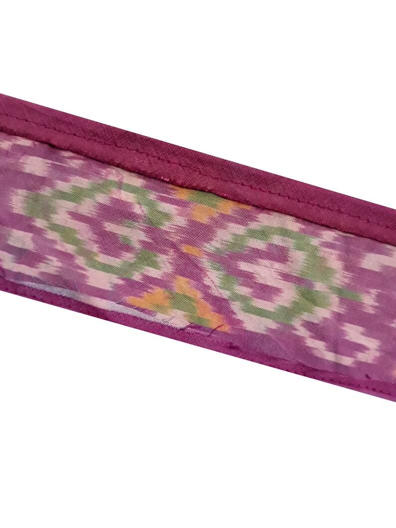 May include: A close-up of a purple fabric with a green and white ikat pattern. The fabric has a pink border.