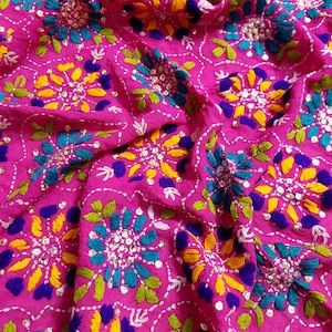 Peut inclure: Gros plan sur un tissu rose vif avec des broderies complexes. Le motif présente des motifs floraux en turquoise, jaune, violet et vert, rehaussés de coutures blanches et de petits sequins argentés. Le tissu est doux et fluide, avec des plis et une texture visibles.