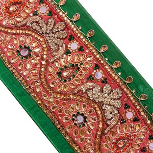 May include: Close-up of a decorative textile with a green border and a central coral panel. The panel is embellished with gold embroidery, sequins, and small, reflective mirror accents. The design features floral and geometric patterns.