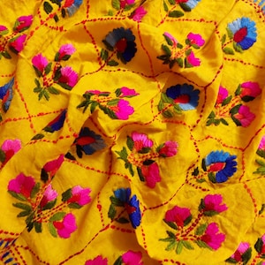 Könnte beinhalten: Leuchtend gelbes Textil mit aufwendiger Stickerei. Das Design zeigt ein sich wiederholendes Muster aus rosa und blauen Blumenmotiven, umrandet mit roten Nähten. Der Stoff ist gerafft und erzeugt ein strukturiertes Aussehen.