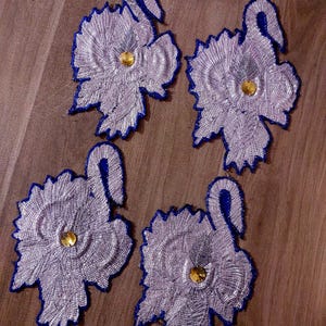 Könnte beinhalten: Vier gestickte Applikationen in Silber und Blau in Form einer stilisierten Blume mit einer goldenen Perle in der Mitte.
