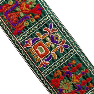 May include: Close-up of a decorative green embroidered ribbon. The ribbon features intricate floral and geometric designs in red, orange, purple, and yellow thread. The edges are outlined with a white, wavy stitch.
