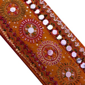 May include: A mustard yellow fabric trim featuring a repeating pattern of circular embroidered motifs with central mirrors. The design includes intricate stitching in pink, green, and white, accented with small metallic sequins and a row of mirrors.