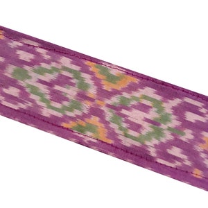 May include: A close-up of a purple fabric with a green, white, and orange ikat pattern. The fabric has a subtle sheen and a soft texture.