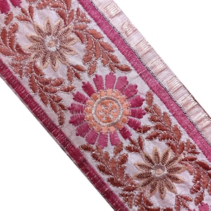 Può includere: Un nastro decorativo in tessuto con un intricato motivo floreale ricamato nei toni del magenta, rame e rosa chiaro. Il design è composto da motivi floreali circolari ripetuti collegati da viticci su una base di colore rosa chiaro.