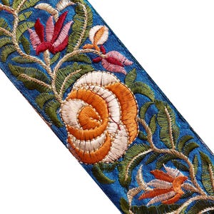 Può includere: Primo piano di un tessuto blu con ricami floreali intricati. Il design presenta fiori arancioni, rosa e bianchi con foglie verdi, tutti cuciti con un'accurata lavorazione del filo. Lo sfondo è blu vibrante.