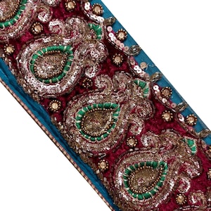 Könnte beinhalten: Eine dekorative Stoffborte mit einer Basis aus tief magenta-farbenem Samt und aufwendiger Stickerei. Das Design zeigt sich wiederholende Paisley-Motive, die mit grünem Garn umrandet, mit Goldperlen gefüllt und mit silbernen Pailletten verziert sind.