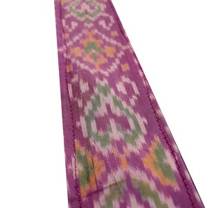 May include: A close-up of a pink fabric with a green, white, and orange ikat pattern. The fabric has a stitched edge.