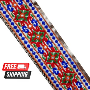 May include: A white fabric trim with a repeating pattern of blue, red, green, and orange embroidery. The trim is decorated with small, round mirrors. The trim is approximately 1.5 inches wide.