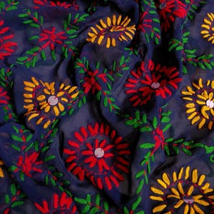 以下が含まれることがあります： 鮮やかな赤、黄、緑の糸で複雑な刺繍が施された、濃紺のシースルー生地。模様は繰り返される花柄で構成されており、それぞれの中心には小さな円形の反射する銀色のスパンコールがあしらわれています。