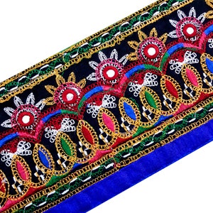 Può includere: Un bordino in tessuto nero con ricami colorati e dettagli a specchio. Il bordino presenta un motivo ripetitivo di fiori, foglie e forme geometriche. Il bordino è rifinito con un tessuto blu.