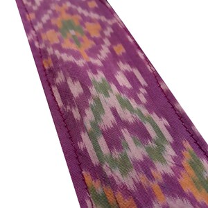 May include: Purple fabric with a green, orange, and white ikat pattern. The fabric has a stitched edge.