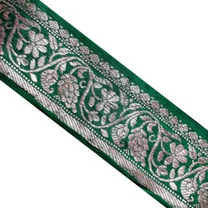 May include: Green fabric trim with a silver floral design. The trim has a repeating pattern of flowers and leaves.