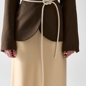 May include: A brown blazer with a cream-colored skirt and a cream-colored rope belt tied at the waist. The blazer has long sleeves and a wrap-style design. The skirt is a solid color and reaches the ankles.