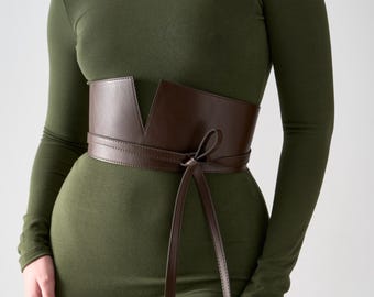 May include: A brown leather waist belt with a V-shaped front design and long tie closure. The belt is worn over a long-sleeved olive green dress. The belt's dimensions are not visible in the image.