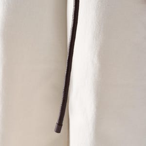 May include: Close-up of a dark brown leather belt with a rounded end. The belt is set against a cream-colored fabric background, creating a contrast in textures and tones. The leather appears smooth and the fabric has a subtle texture.