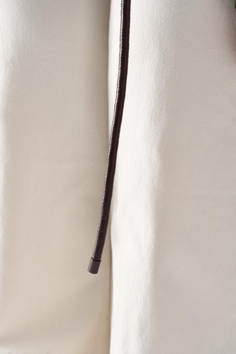 May include: Close-up of a dark brown leather belt with a rounded end. The belt is set against a cream-colored fabric background, creating a contrast in textures and tones. The leather appears smooth and the fabric has a subtle texture.