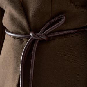 May include: Close-up of a brown blazer with a matching leather belt tied in a knot. The blazer is made of a textured fabric and has a classic design. The belt adds a stylish accent to the garment.