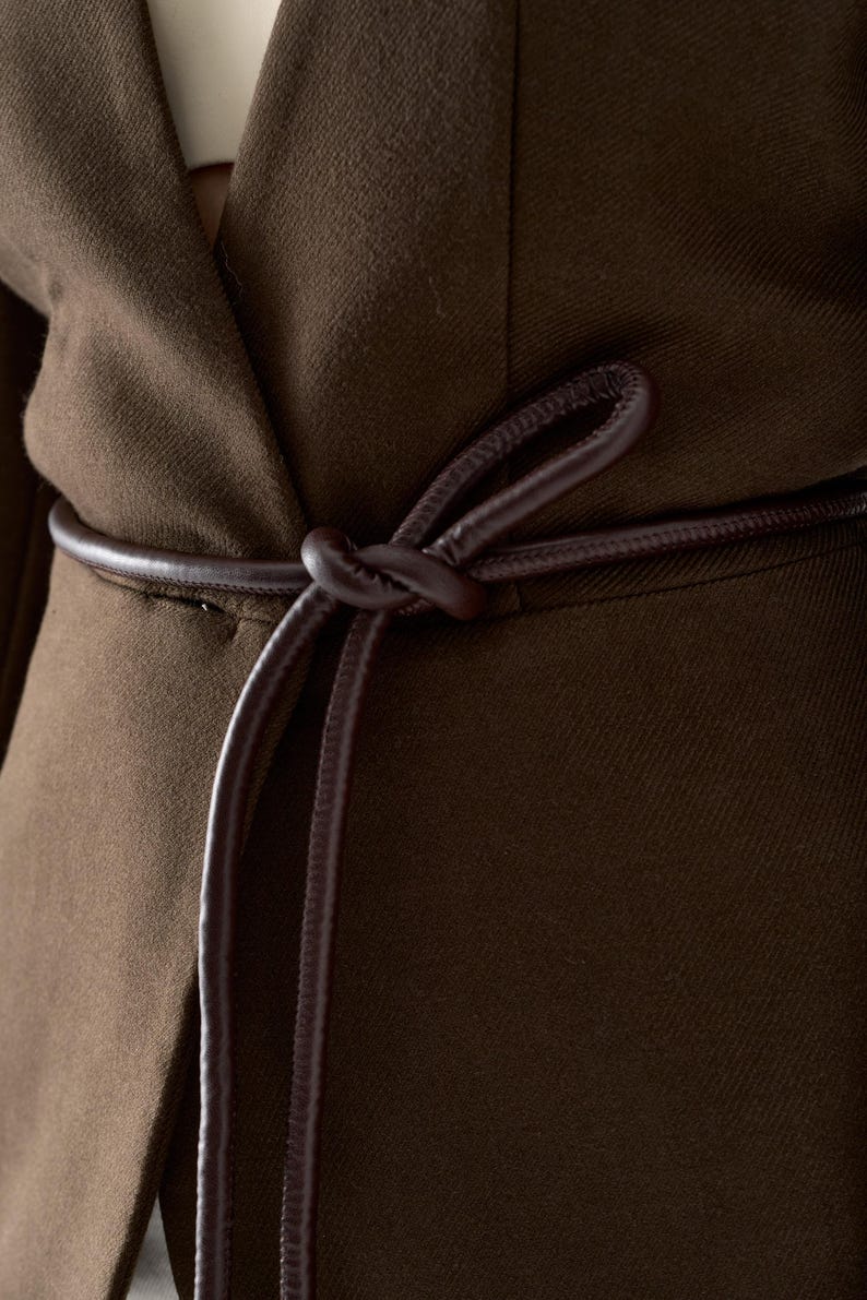 May include: Close-up of a brown blazer with a matching leather belt tied in a knot. The blazer is made of a textured fabric and has a classic design. The belt adds a stylish accent to the garment.