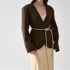 May include: A dark brown blazer with a V-neck design and a cream-colored rope belt. The blazer is paired with a beige dress. The outfit is simple and elegant, suitable for a variety of occasions.