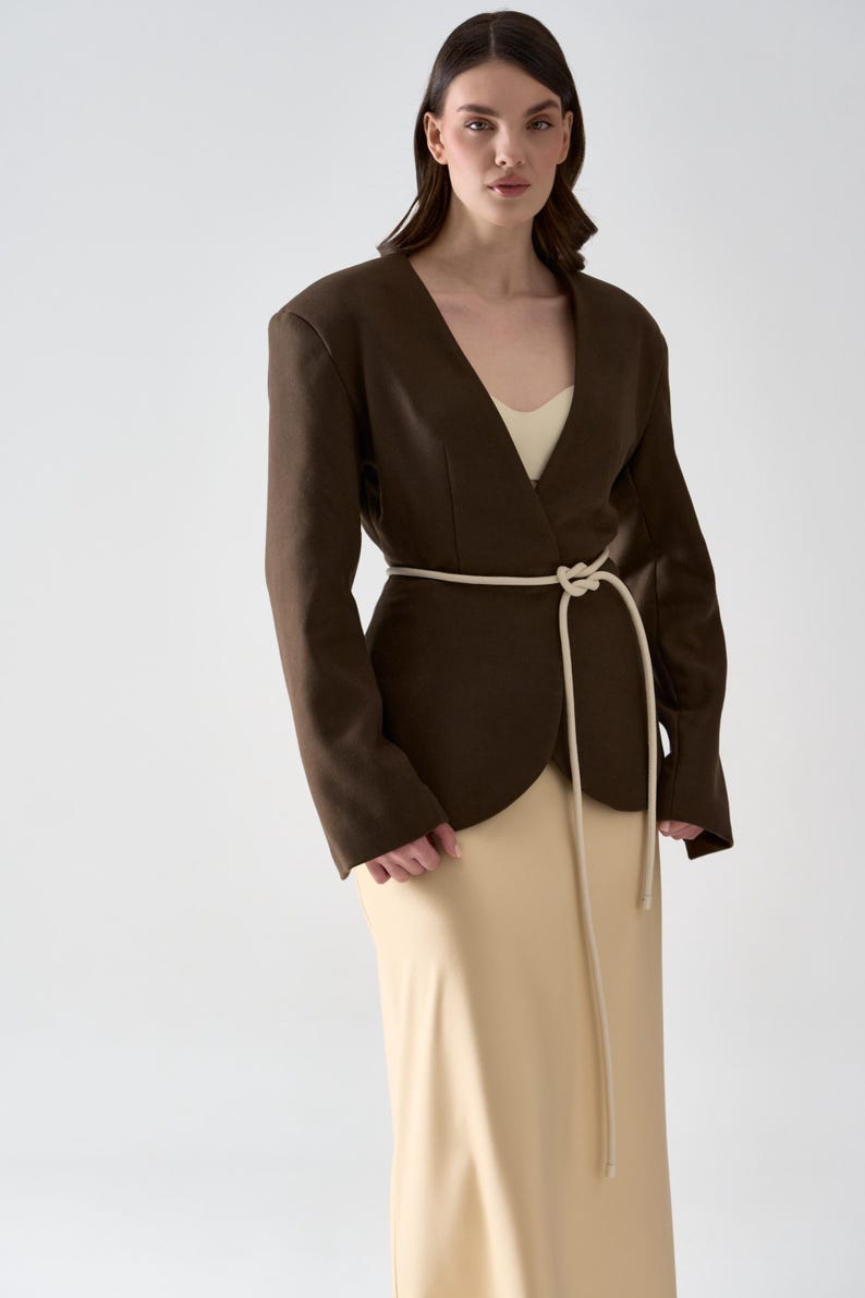 May include: A dark brown blazer with a V-neck design and a cream-colored rope belt. The blazer is paired with a beige dress. The outfit is simple and elegant, suitable for a variety of occasions.