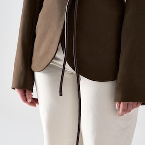 May include: A dark olive green blazer with a matching brown belt tied at the waist. The blazer has long sleeves and a tailored fit. The pants are off-white. The outfit is simple and elegant.