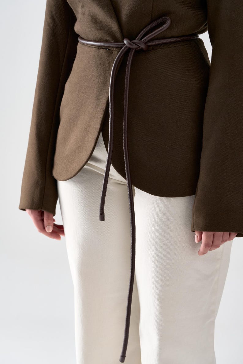 May include: A dark olive green blazer with a matching brown belt tied at the waist. The blazer has long sleeves and a tailored fit. The pants are off-white. The outfit is simple and elegant.