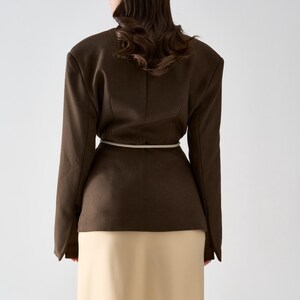May include: A dark brown blazer with a cream-colored belt and a long, cream-colored skirt. The blazer has structured shoulders and long sleeves. The model is facing away from the camera.