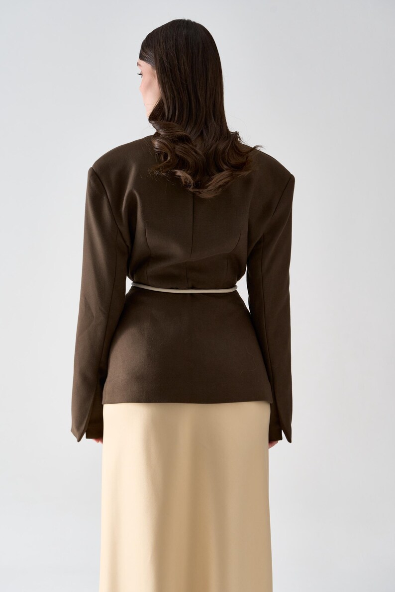 May include: A dark brown blazer with a cream-colored belt and a long, cream-colored skirt. The blazer has structured shoulders and long sleeves. The model is facing away from the camera.