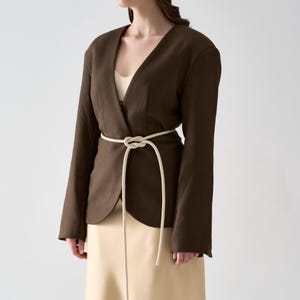 May include: A dark brown blazer with a V-neck design and long sleeves. The blazer is cinched at the waist with a cream-colored rope belt. The model is wearing a cream-colored top and a beige skirt.