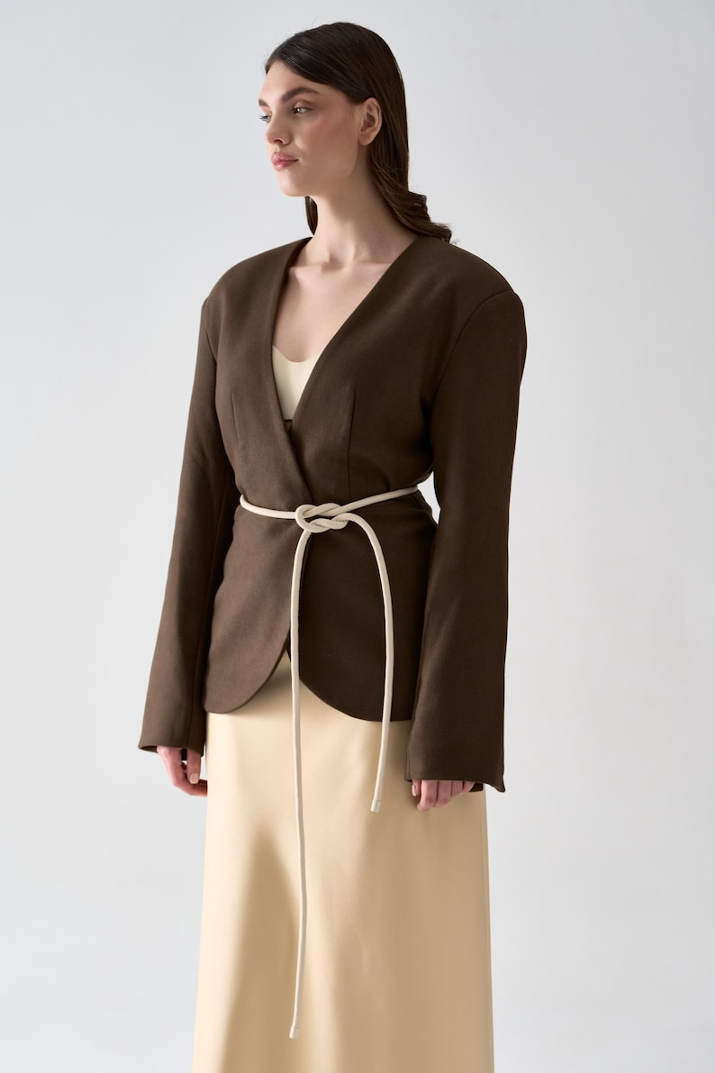 May include: A dark brown blazer with a V-neck design and long sleeves. The blazer is cinched at the waist with a cream-colored rope belt. The model is wearing a cream-colored top and a beige skirt.