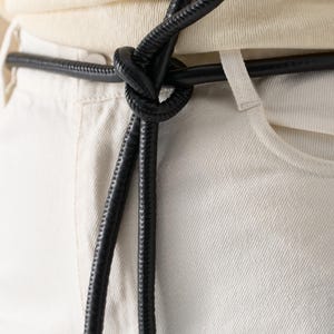 May include: Close-up of a black leather belt tied in a knot. The belt is worn over light-colored pants and a cream-colored top. The belt's texture is visible, and the knot is the focal point.