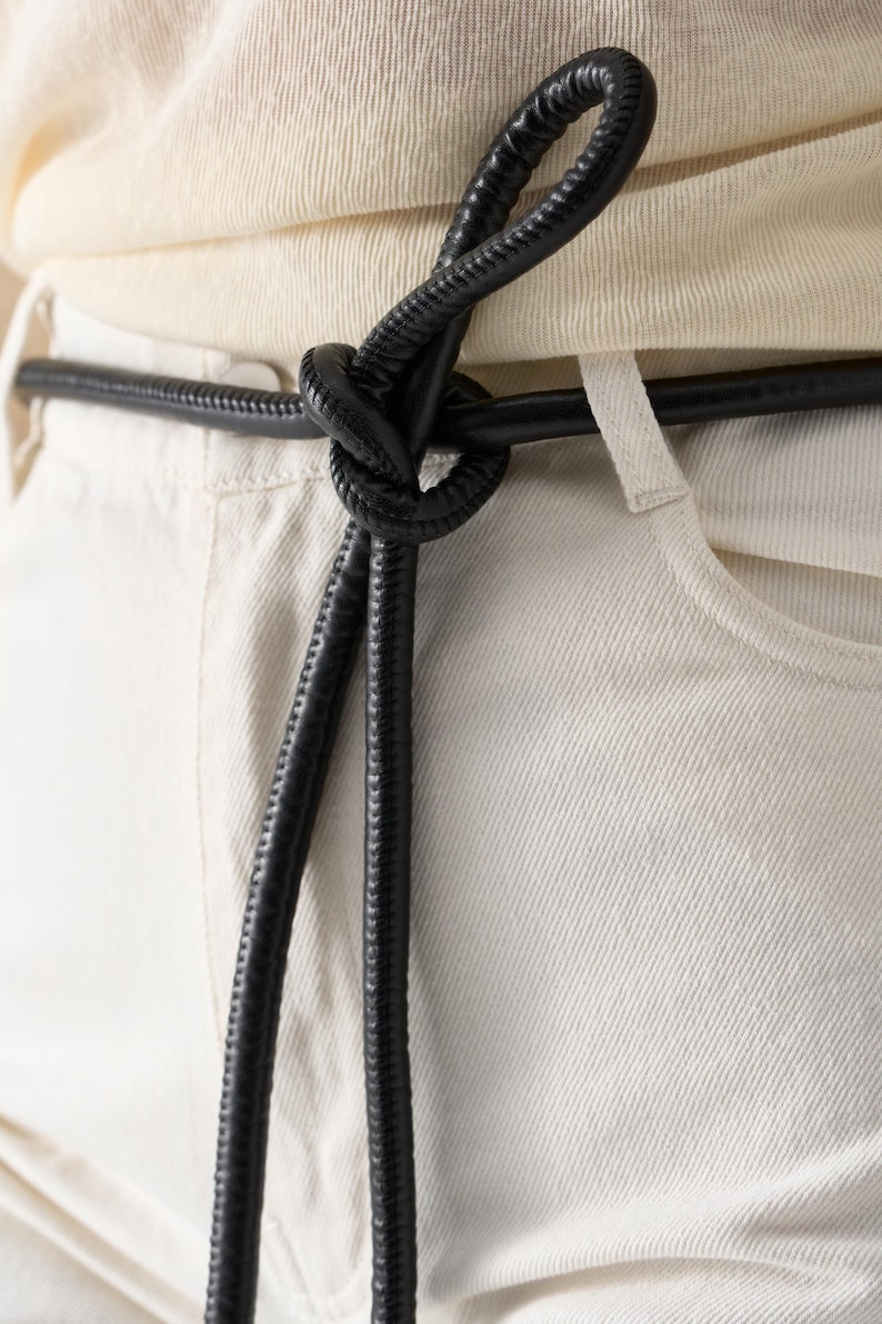 May include: Close-up of a black leather belt tied in a knot. The belt is worn over light-colored pants and a cream-colored top. The belt's texture is visible, and the knot is the focal point.