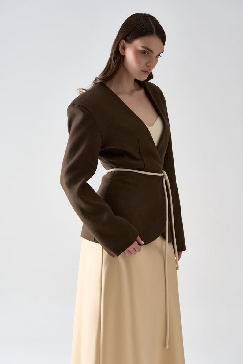 May include: A dark brown blazer with a V-neck design and a cream-colored belt. The blazer is paired with a long, beige skirt. The outfit is simple and elegant, suitable for various occasions.