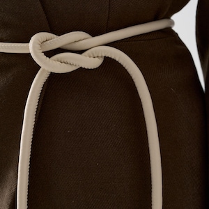 Könnte beinhalten: Nahaufnahme eines dunkelbraunen Kleidungsstücks mit einem cremefarbenen Gürtel, der zu einem Knoten gebunden ist. Der Gürtel besteht aus einem weichen, abgerundeten Material. Das Kleidungsstück hat ein strukturiertes Aussehen.