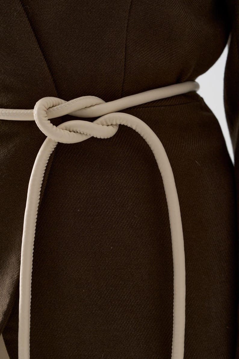 May include: Close-up of a dark brown garment with a cream-colored belt tied in a knot. The belt is made of a soft, rounded material. The garment has a textured appearance.