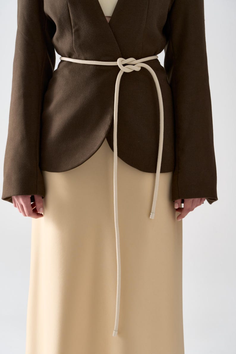 May include: A brown blazer with a cream-colored skirt and a cream-colored rope belt tied at the waist. The blazer has long sleeves and a wrap-style design. The skirt is a solid color and reaches the ankles.
