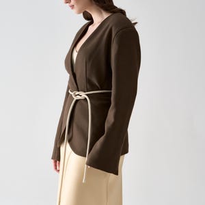 May include: A dark brown blazer with a V-neck and long sleeves, cinched at the waist with a cream-colored rope belt. The blazer is paired with a cream-colored skirt. The outfit is simple and elegant.
