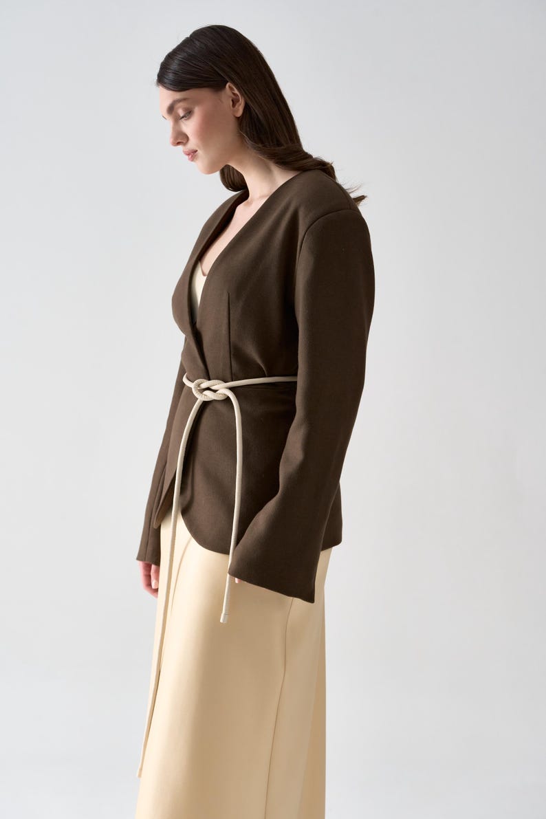 May include: A dark brown blazer with a V-neck and long sleeves, cinched at the waist with a cream-colored rope belt. The blazer is paired with a cream-colored skirt. The outfit is simple and elegant.