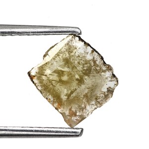 May include: A raw, pale yellow diamond held by tweezers against a white backdrop. The diamond is roughly square with uneven edges and a translucent quality, revealing internal patterns and textures.